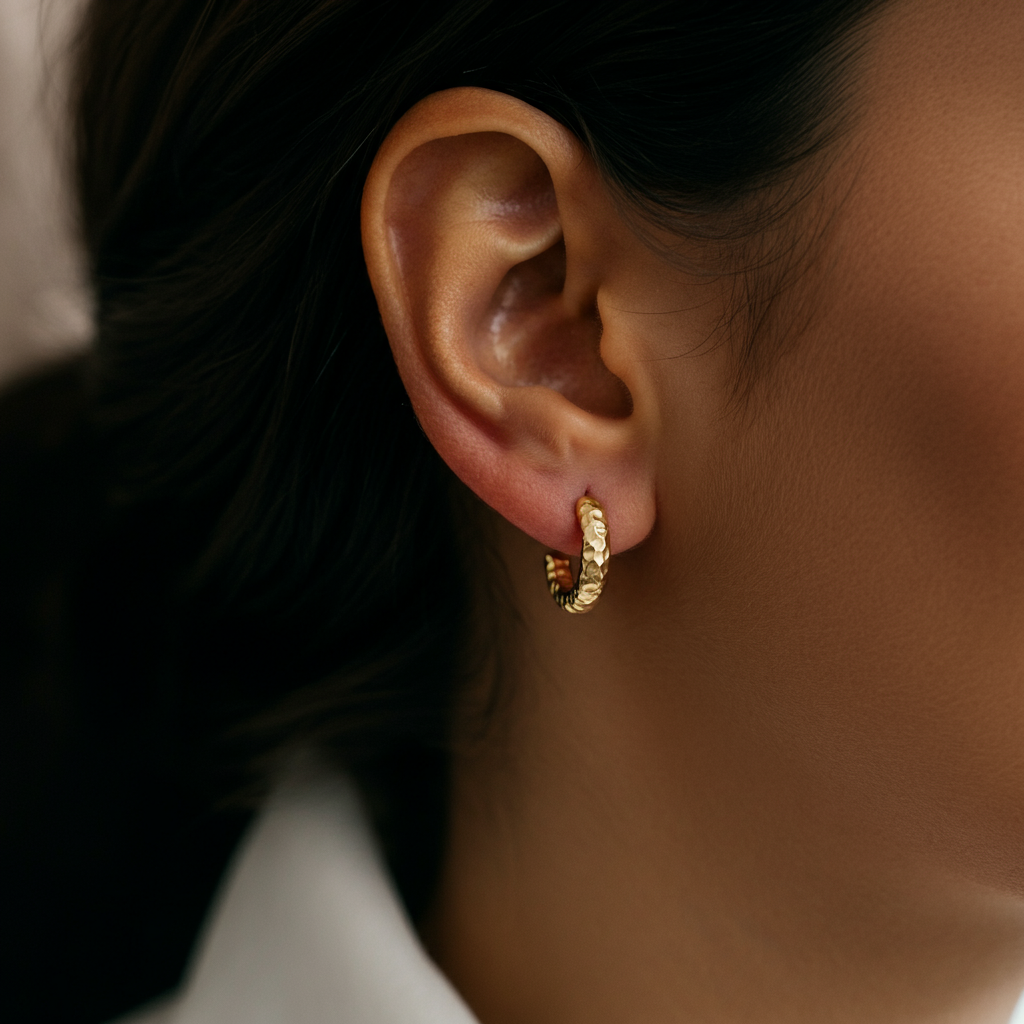 Close-up shot of a woman's ear wearing small, gold hoop earrings. The lighting is soft and diffused, accentuating the texture of the gold. The background is blurred to keep the focus on the earrings.